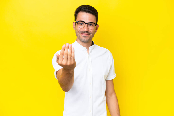 Young handsome man over isolated yellow background inviting to come with hand. Happy that you came - Photo, Image