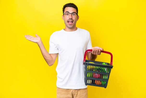 Jovem homem bonito segurando uma cesta de compras cheia de comida sobre fundo amarelo isolado com expressão facial chocada - Foto, Imagem