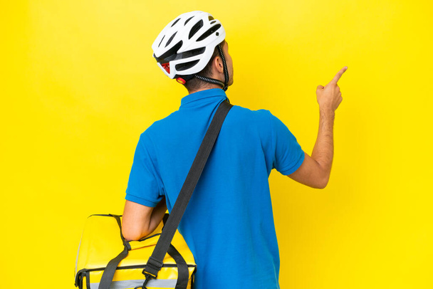 Young handsome man with thermal backpack over isolated yellow background pointing back with the index finger - Фото, изображение