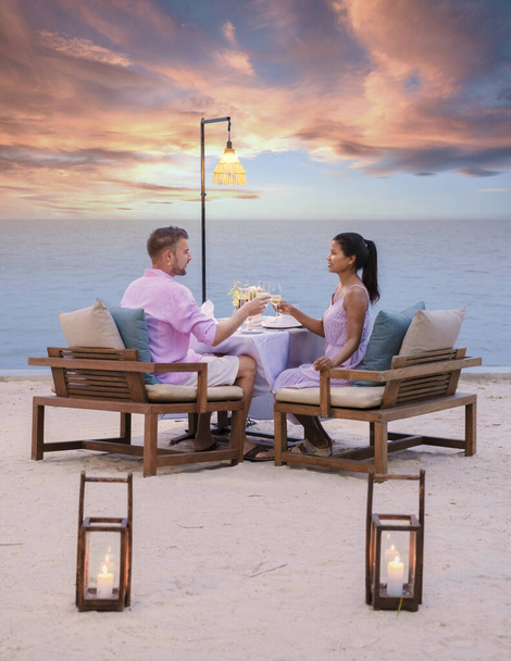 夕方にはビーチでロマンチックなディナーを楽しんでいる男女のカップル。アジア系の女性と白人男性がフアイン・タイのビーチで夕食をとる - 写真・画像