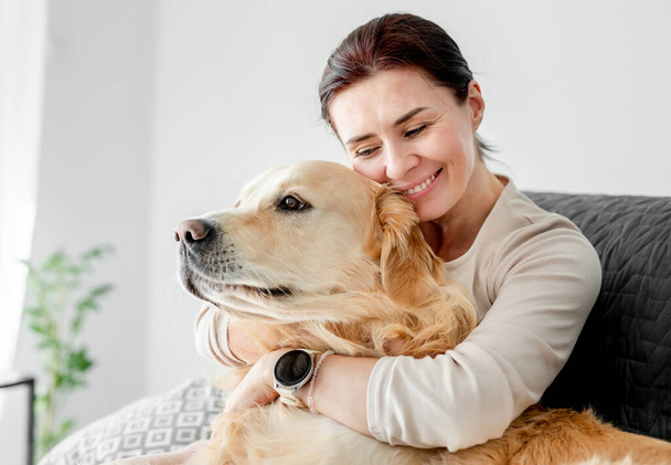 Girl hugging golden retriever dog at home and smiling. Young woman petting doggy with love - Foto, Imagem