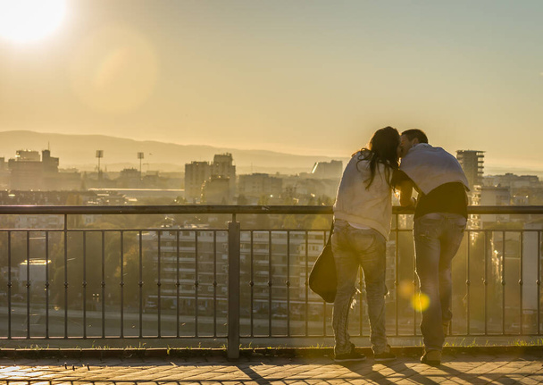 Ένα ερωτευμένο ζευγάρι, να στέκεται ακουμπισμένο σε ένα μεταλλικό φράχτη. Ένα ερωτευμένο ζευγάρι, φωτισμένο από τον απογευματινό ήλιο, ακουμπά σε ένα μεταλλικό φράχτη.. - Φωτογραφία, εικόνα