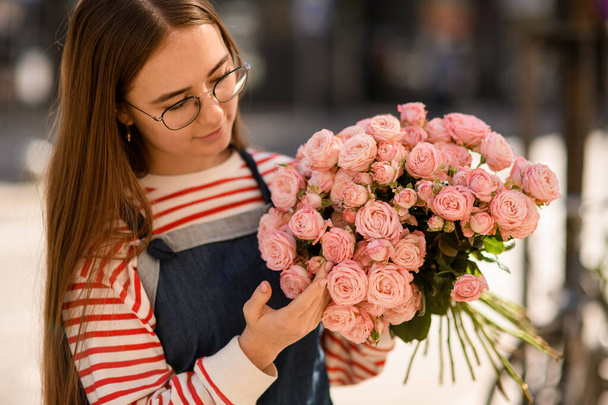 portrait of pretty young woman in glasses with long hair holding bouquet of fresh light pink roses in her hands - Fotó, kép