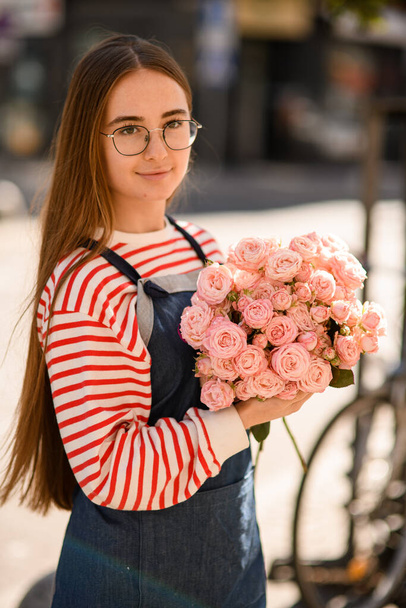 portrait of beautiful young woman in glasses with long hair holding bouquet of fresh light pink roses in her hands - Photo, image