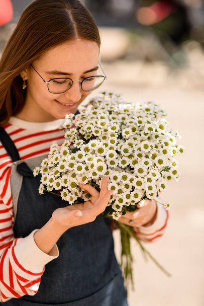 Handsome young smiling woman with bouquet of fresh white small daisies. Blurred background. Close-up - Фото, изображение