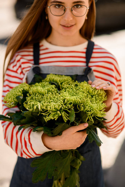 mujer joven en gafas con pelo largo sostiene ramo de flores de crisantemo verde fresco en sus manos - Foto, Imagen