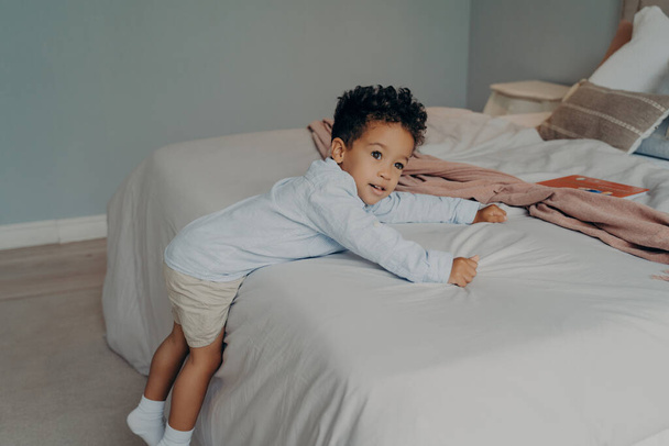 Sweet little afro american kid boy in stylish cotton clothes trying to climb on big white soft bed, holding onto bedsheet while playing in bright bedroom at home, preschool child spending leisure time - Photo, Image