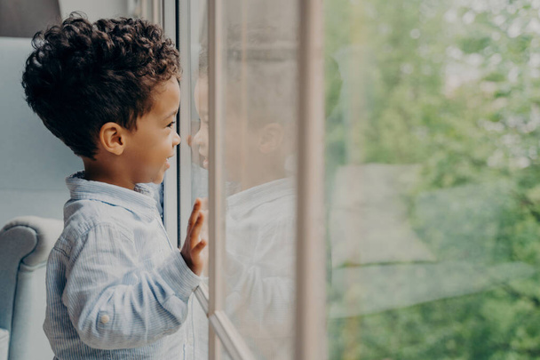 Vista lateral de un niño afroamericano encantador esperando a mamá, vestido con camisa azul, mirando por la ventana a través de su reflejo apoyado en el vidrio con las manos y sonriendo. Niños tiempo libre interior - Foto, imagen
