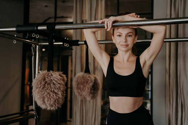 Young happy ginger woman in black sportswear taking break on pilates machine between trainings in gym,joyfully looking forward smiling on camera, healthy lifestyle and sport indoors concept - Foto, Imagem