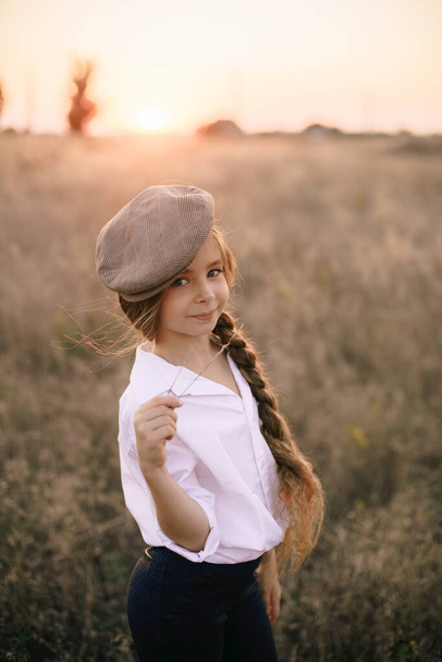cute little girl with a long braid of blond hair in a white shirt and a men's vintage cap at sunset in a summer field. - Фото, изображение