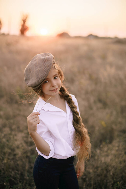 cute little girl with a long braid of blond hair in a white shirt and a men's vintage cap at sunset in a summer field. - Foto, Bild