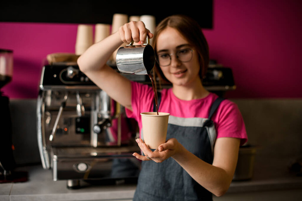 Selective focus on steel jug in the hand of female barista from which coffee is poured into paper cup on blurred background - Fotoğraf, Görsel