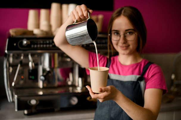 view of steel jug in the hand of female barista from which milk is poured into paper cup on blurred background - Фото, изображение