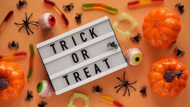 Pumpkins, cauldrons, pots, worms, jaws, spiders, eye-shaped candies and a text board with the inscription TRICK OR TREAT on a orange background, top view. A collection of items for a Halloween party - Фото, зображення