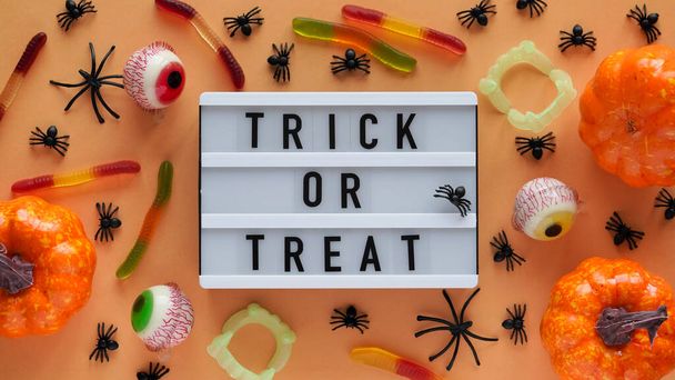 Pumpkins, cauldrons, pots, worms, jaws, spiders, eye-shaped candies and a text board with the inscription TRICK OR TREAT on a orange background, top view. A collection of items for a Halloween party - Photo, Image