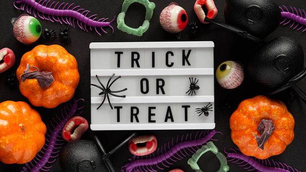 Pumpkins, cauldrons, pots, worms, jaws, spiders, eye-shaped candies and a text board with the inscription TRICK OR TREAT on a black background, top view. A collection of items for a Halloween party - Photo, Image