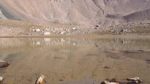 Läpinäkyvä vuoristojärvi kivien ympäröimänä. Vesi on kuin peili, joka heijastaa kiviä ja rinteitä. Valo väreilee veden pinnalla. Pohja on näkyvissä. Siellä on isoja kiviä - Materiaali, video