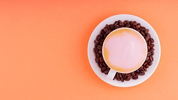 Cup of coffee with pink foam on a coral background. Coffee cup and roasted coffee beans on a saucer. Top view. Copy space - 写真・画像