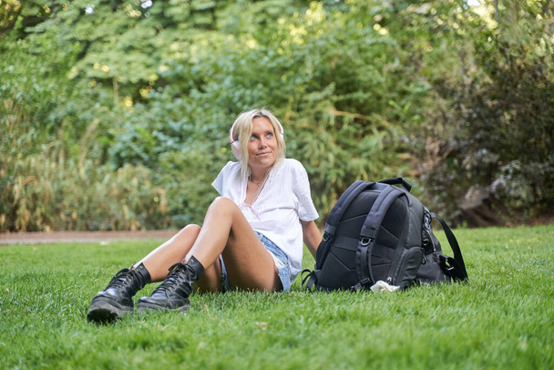 Νεαρή γυναίκα που απολαμβάνει να ακούει μουσική με ακουστικά ενώ χαλαρώνει κάθεται στο γρασίδι σε ένα πάρκο. Τεχνολογία και ελεύθερος χρόνος έννοια. - Φωτογραφία, εικόνα