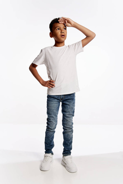 Looking up suspiciously. Full-length portrait of african little boy in white tee expressing emotions isolated over white background. Kids emotions, news, challenges, childhood, education concept - Foto, Imagen