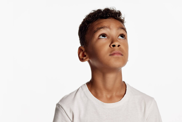 Dreaming. Cute african little boy wearing white t-shirt looks up in surprise isolated over white background. Kids emotions, goals, dreams, hope concept. Copy space for ad - Foto, Imagem