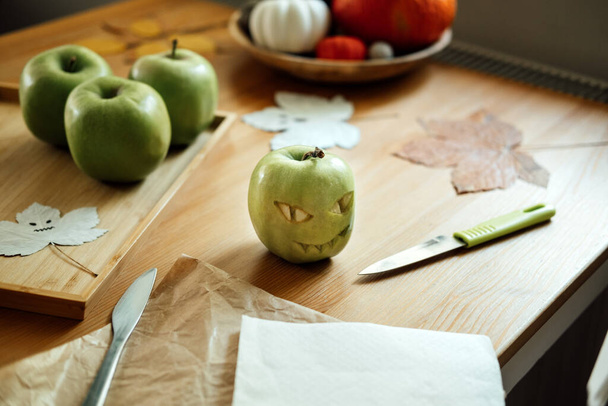 Фруктовые идеи на Хэллоуин. Зеленое яблоко на Хэллоуин с вырезанным лицом на деревянном столе дома. Здоровые Хэллоуинские закуски. - Фото, изображение