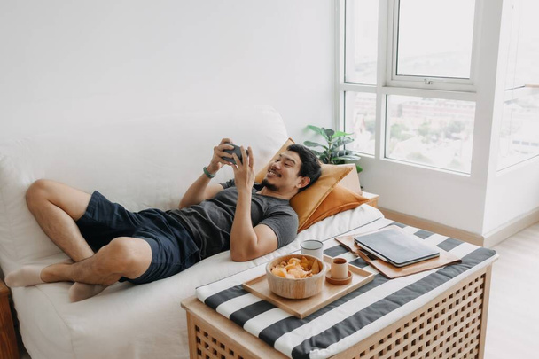 彼は家から仕事を持っている必要がありながら、彼はモバイルゲームをプレイ怠惰な幸せなアジアの男. - 写真・画像