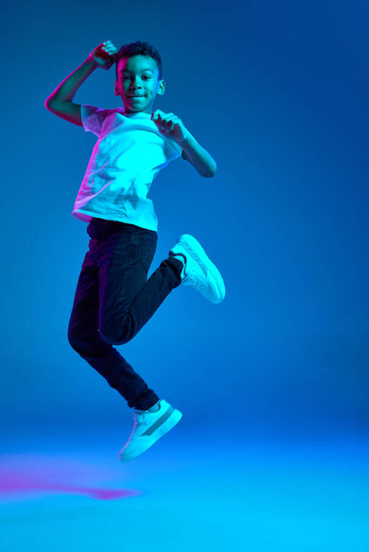 Jumping, having fun. Studio shot of emotional little boy, kid expressing different emotions isolated over blue background in neon light. Education, childhood, modern lifestyle - Φωτογραφία, εικόνα