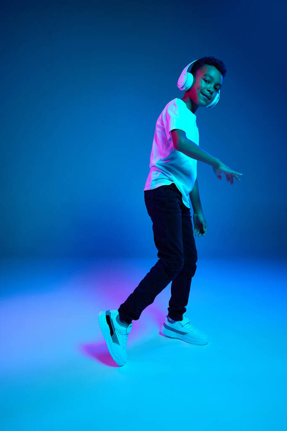 Dance. Little cheerful happy boy 6-7 years old wearing white t-shirt and headphones listen to music isolated on blue background in neon light. Concept of childhood, music, dance. Looks joyfull - Zdjęcie, obraz