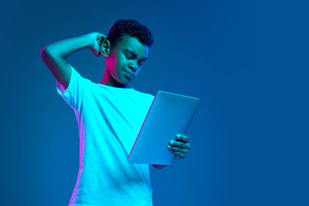 Remote education. Serious little boy, kid in white t-shirt looking at tablet screen isolated over blue background in neon light. Studying, gadgets, childhood, modern lifestyle - Foto, imagen