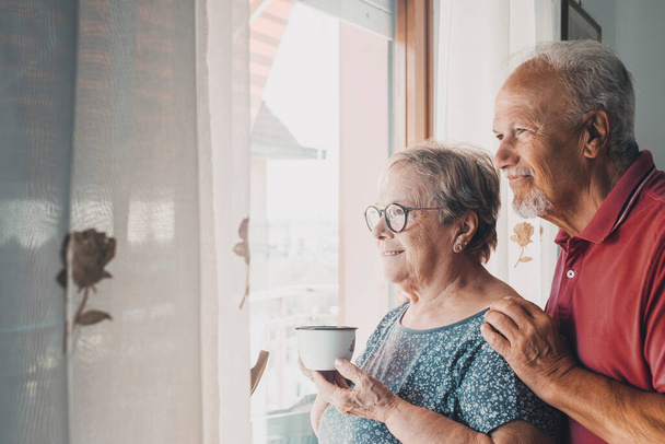 Ein älteres Ehepaar blickt gemeinsam aus dem Fenster auf wartende Eltern. Großväter in Freizeitaktivitäten in Innenräumen. Liebe zwischen alten, reifen Menschen.  - Foto, Bild