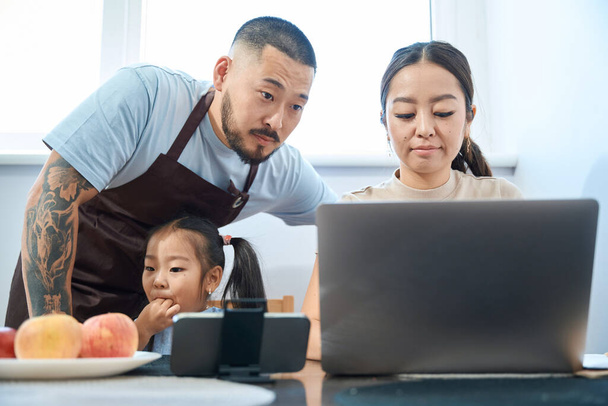 Ázsiai férj fölé hajolt a felesége laptop, lányuk lófarkú ül az étkező asztal mellett - Fotó, kép