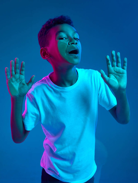 Close-up of little boy leaning against transparent glass by cheek isolated on blue background in neon light. Funny cute face. Concept of emotions, childhood, facial expression. Copy space for ad - Foto, Imagen