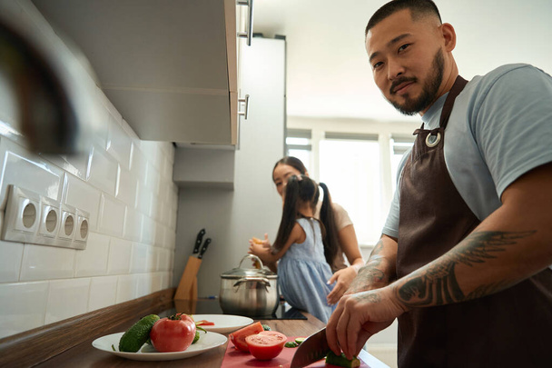 台所のアジアの男は野菜を切り、彼の妻と娘の隣に調理するのを助ける - 写真・画像