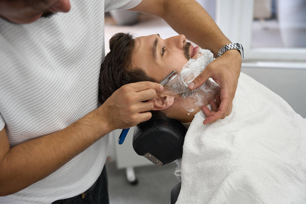 Cropped photo of experienced barber using straight razor while shaving face of his client - Φωτογραφία, εικόνα