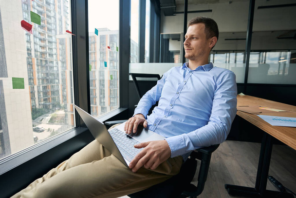 Zamyšlený zaměstnanec společnosti sedí v křesle s notebookem na klíně a dívá se oknem - Fotografie, Obrázek