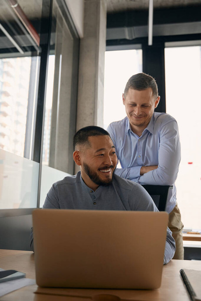 Smiling company employee leaning on chair backrest of his cheerful colleague seated at laptop - Photo, image