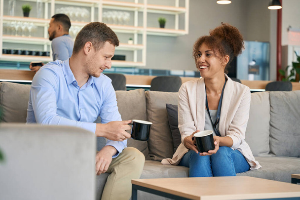 Jongeman en vrolijke vrouw zitten op de bank met elkaar te praten tijdens de koffiepauze - Foto, afbeelding
