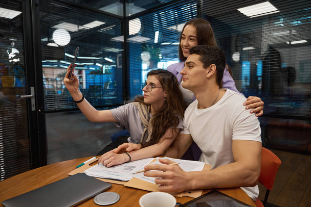 Colleghi felici tre di loro sono seduti a una scrivania in ufficio mentre uno dei dipendenti ha allungato la mano in avanti per un selfie - Foto, immagini