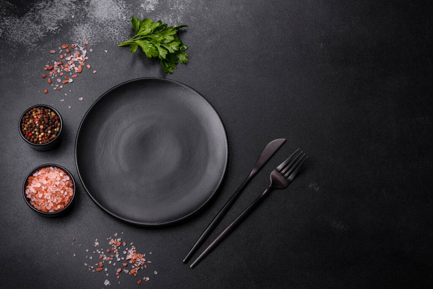An empty black plate on a dark concrete background with spices, herbs and cutlery. Home dinner cooking - Фото, изображение