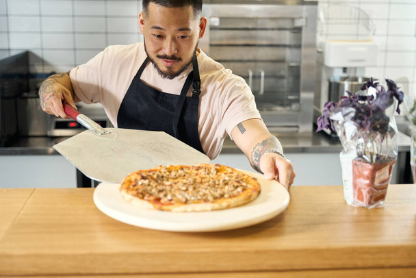 Cocinero profesional mirando enfocado mientras pone una pizza recién cocinada en el plato - Foto, Imagen