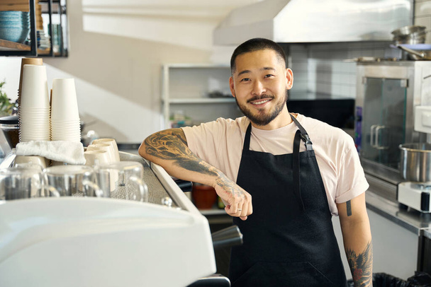 Foto en la cintura de un joven barista orgulloso apoyado en una cafetera y sonriendo a la cámara - Foto, Imagen
