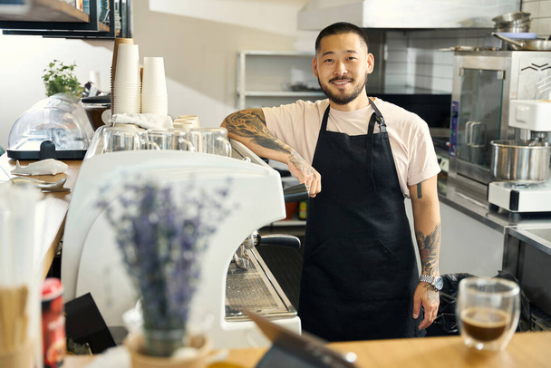 Симпатичный мужчина в фартуке позирует рядом с кофеваркой в современном кафе - Фото, изображение