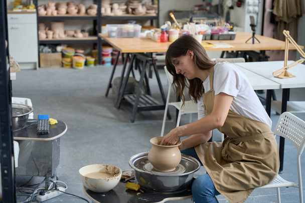 Yaratıcı bir atölyede çömlekçi kadın çömlekçi vazosuyla çömlekçi çarkında çalışıyor. - Fotoğraf, Görsel