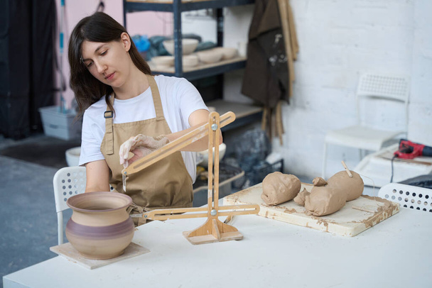 La mujer se sienta en su escritorio y trabaja con una brújula de alfarero, junto a una jarra, trozos de arcilla, herramientas - Foto, imagen