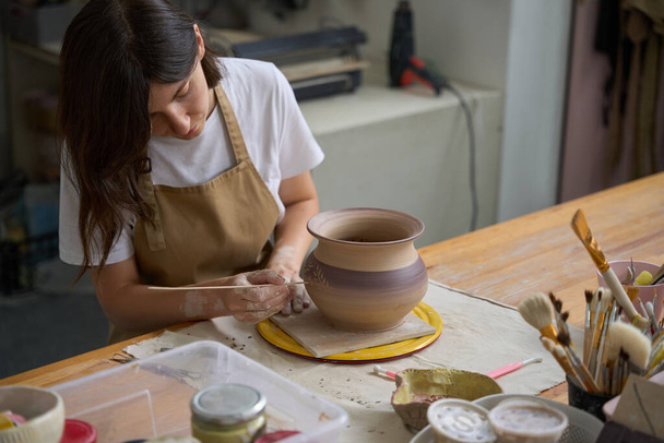 Artigiana in un laboratorio di ceramica si siede a un tavolo e lavora con una spatola su un vaso di argilla - Foto, immagini