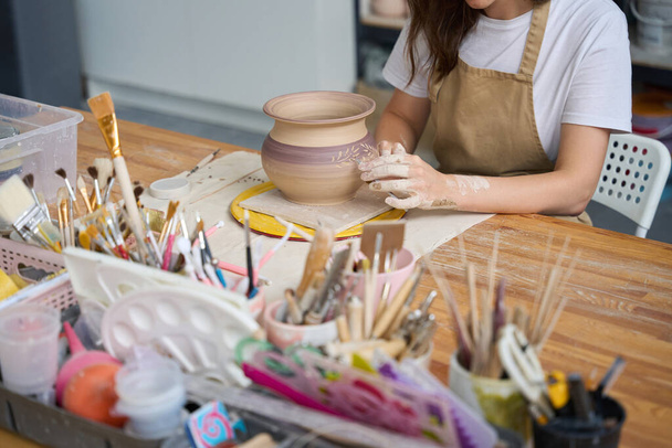 Craftswoman sits at a simple wooden table and applies a decorative floral pattern to a clay pot - Photo, Image