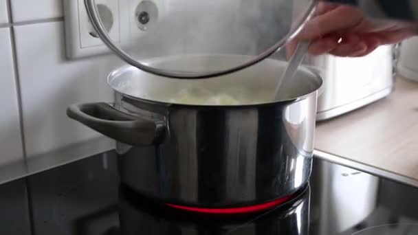 Vrouw controleren kokende aardappelen in warm water in zilveren pot op keramische kachel heffen deksel met stoomstroom toont authentieke koken thuis om gezonde maaltijd te bereiden met het koken van aardappelen op keramische kachel - Video