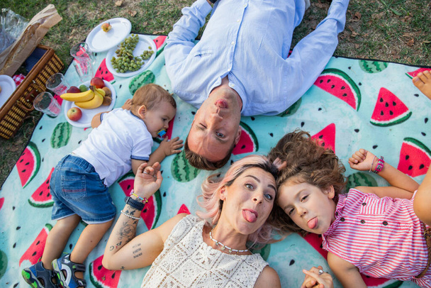 Beautiful family with small children at the park lying on the ground having picnic doing funny faces. - Photo, Image