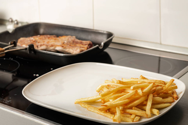 Plate of French fries next to a grill with a steak cooking. - Φωτογραφία, εικόνα